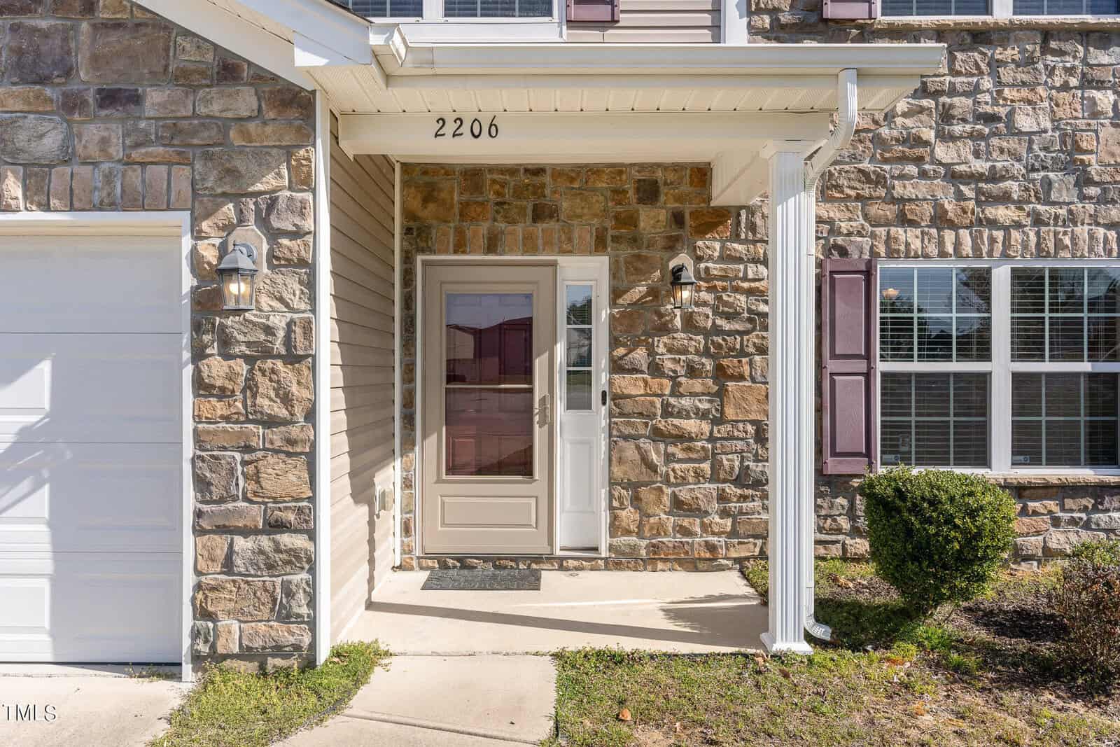Front view of a house featuring a covered entryway with stone and siding exterior. The house number "2206" is above the door, and there is a lantern on the wall. A small bush and grass are near the pathway leading to the door.