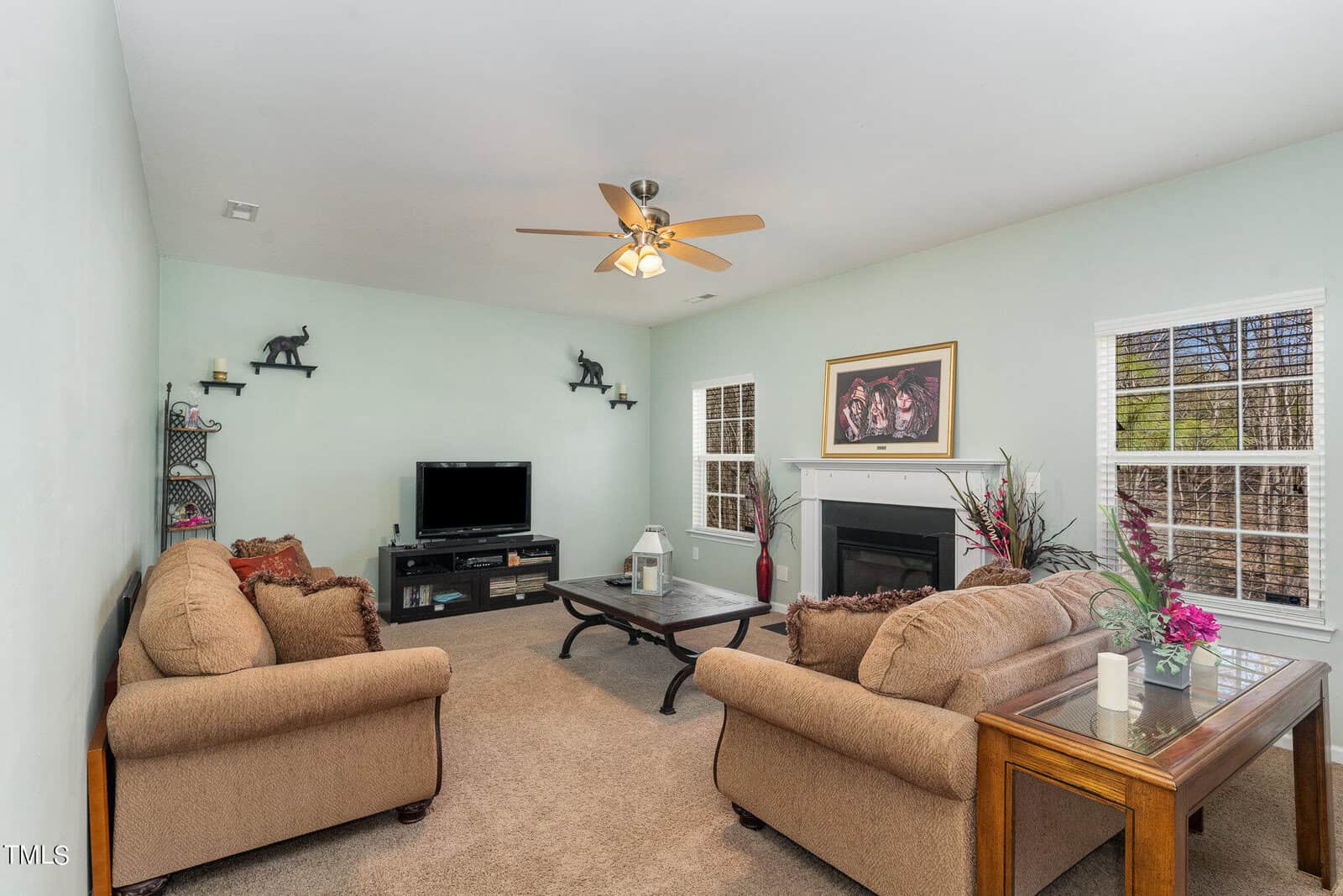 A cozy living room with pale green walls, featuring two beige sofas, a wooden coffee table, and a TV on a stand. A ceiling fan hangs above. The room has a fireplace with a framed picture above it and windows letting in natural light.
