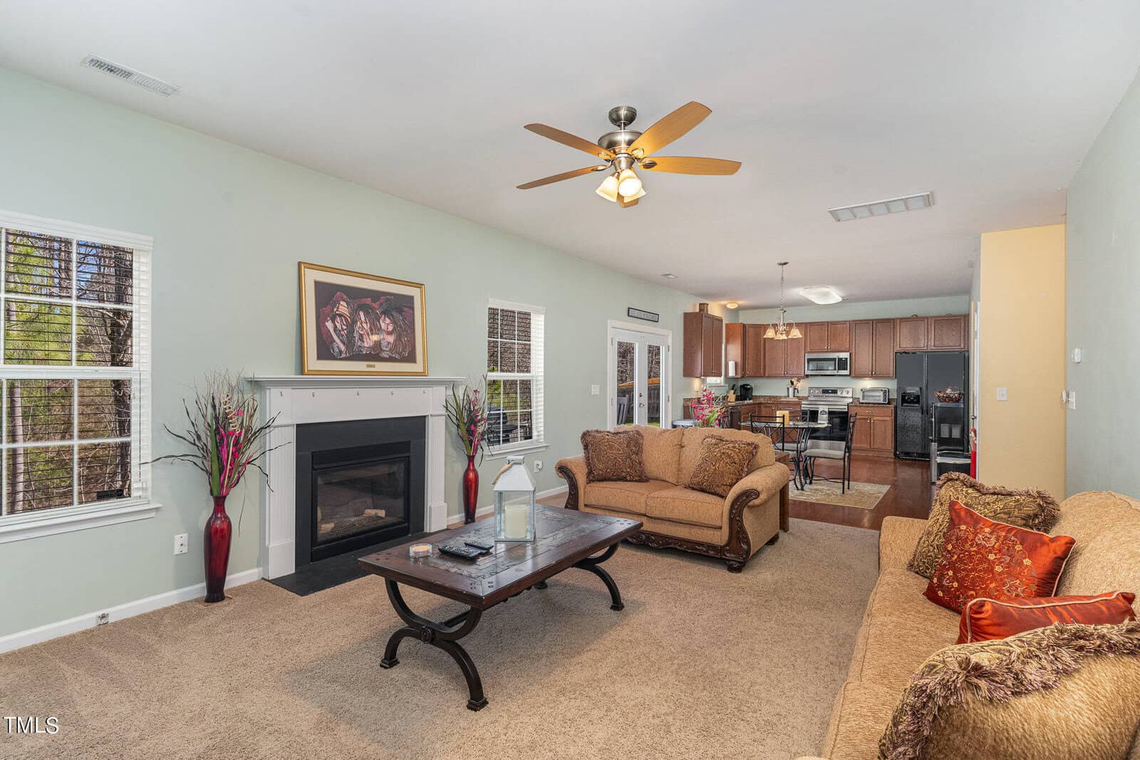 A cozy living room with beige sofas, a coffee table, and a fireplace on a light green wall. The room connects to a dining area and a kitchen with wooden cabinets. A ceiling fan hangs above, and a painting adorns the wall between two windows.