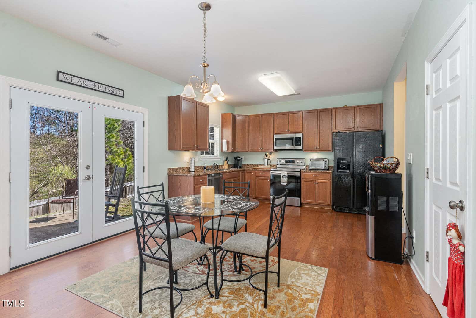 A kitchen and dining area with wood flooring. The dining table has four iron chairs with beige cushions. The kitchen features wooden cabinets and black appliances. A door with glass panels leads to an outdoor deck with tall trees visible.