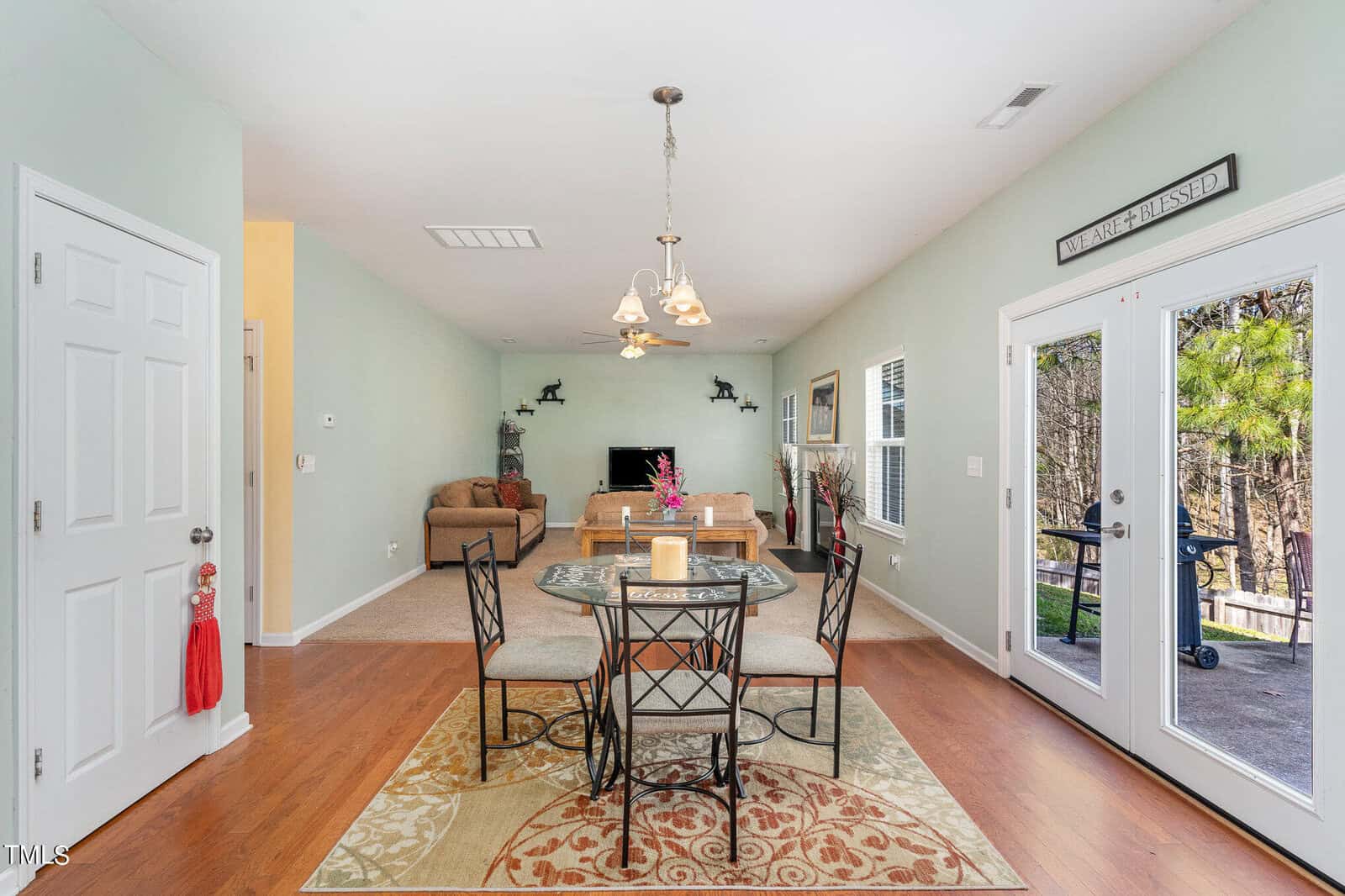 A dining area with a round glass table and four chairs on a patterned rug. The room has light green walls, wood flooring, and large windows. A chandelier hangs above, and a living space with a TV is visible in the background.
