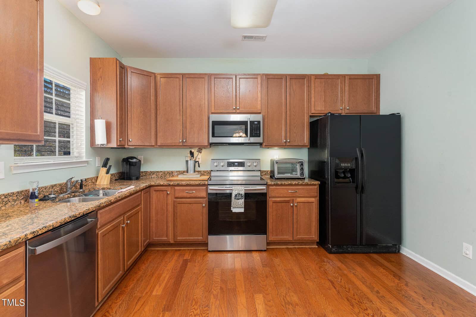 A kitchen with wooden cabinets, black appliances including a refrigerator and an oven, a dishwasher, and a microwave. The countertop is granite, and there is a window above the sink allowing natural light in. The floor is hardwood.