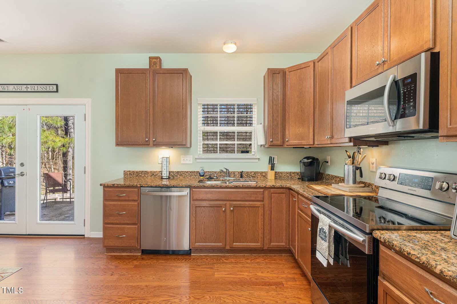 A cozy kitchen with wooden cabinets, stainless steel appliances, and a granite countertop. A window above the sink lets in natural light, and a glass door opens to an outdoor patio. The walls are light green, complementing the warm wood tones.