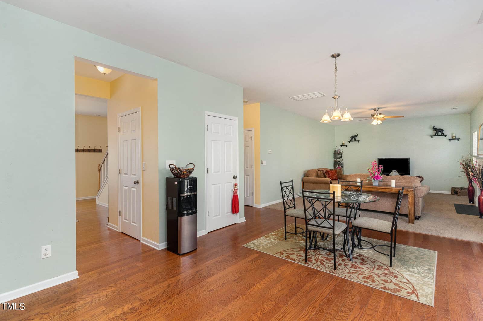 Interior of an open-plan living and dining area with light green walls and hardwood flooring. Features a round dining table with chairs, a ceiling fan, and a beige sectional sofa. A TV is mounted on the wall, and there's a water dispenser near a hallway.