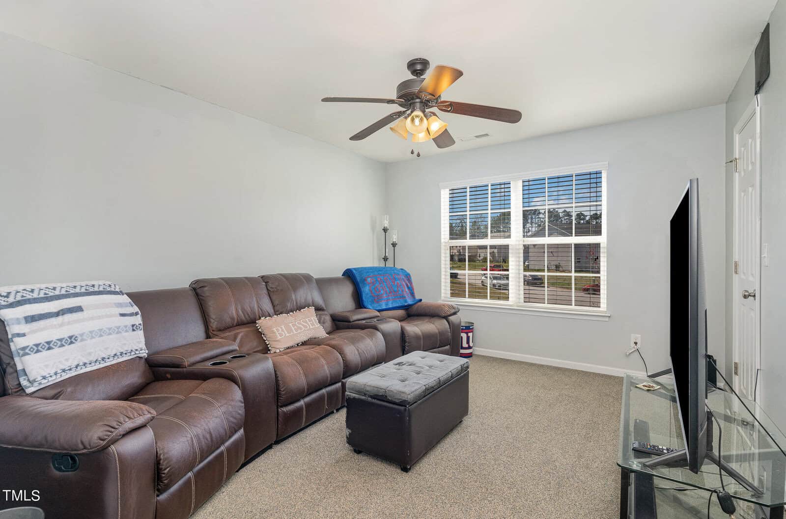 A cozy living room with a brown leather sectional sofa and blue throw blankets. A ceiling fan hangs overhead. A television on a stand is placed opposite the sofa. A window lets in natural light, and a black ottoman sits in front of the sofa.