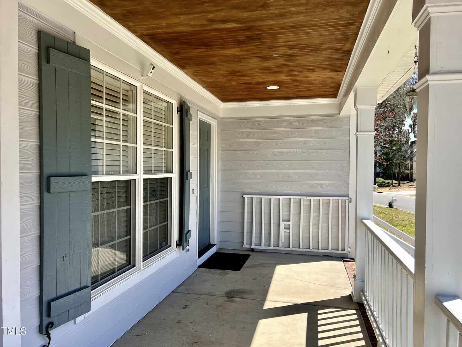 A house porch with light blue siding and a wooden ceiling. There are two windows with blue shutters on one side and a closed door on the other. A small railing and white pillars border the porch, leading to a view of the street.
