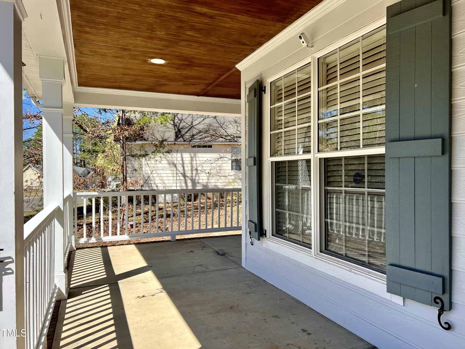 A sunlit porch with a wooden ceiling and a concrete floor features white railings and a large window with green shutters. A shed and trees are visible in the background under a clear blue sky.
