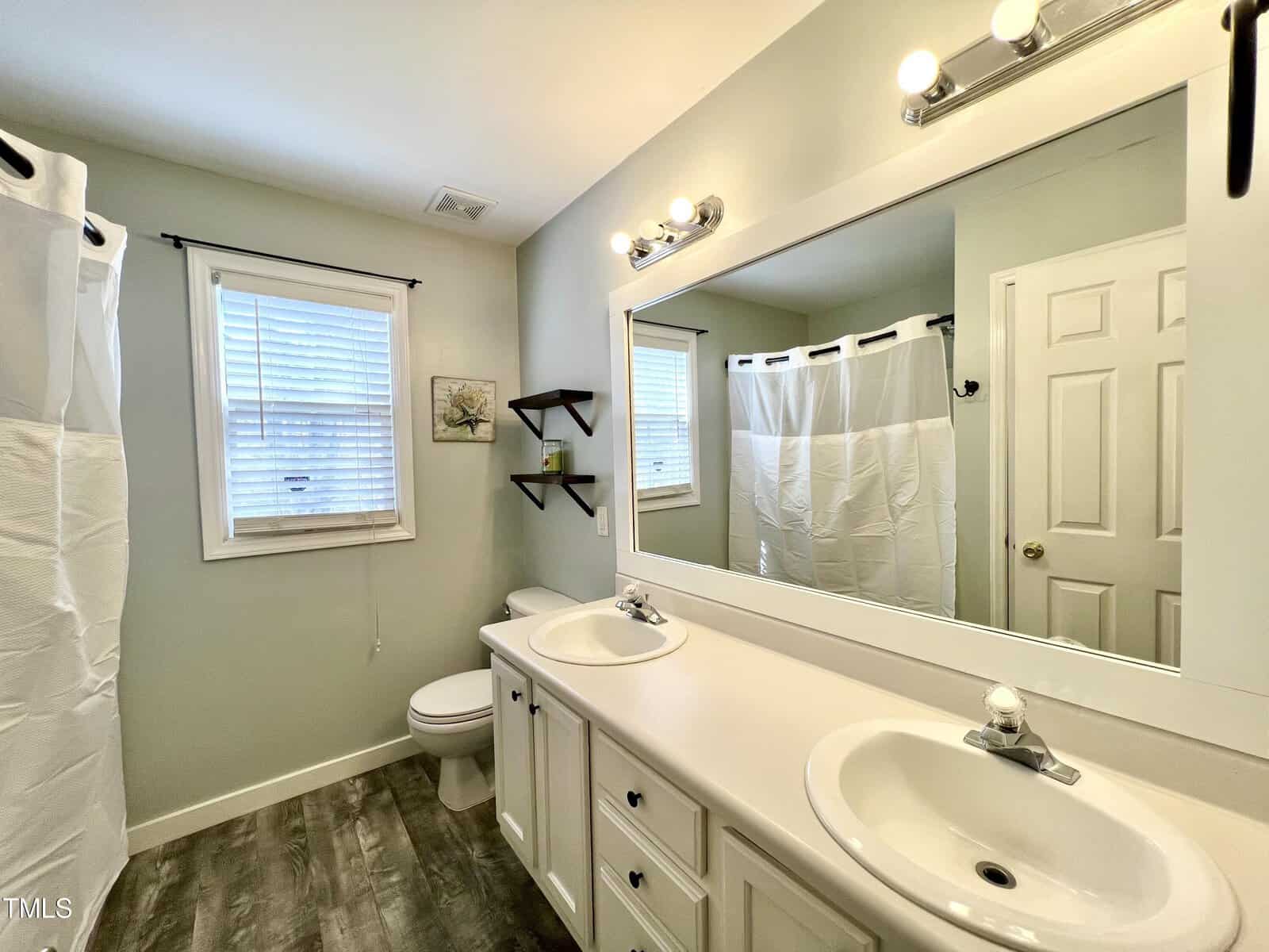 A clean, white bathroom with a double sink vanity and a large mirror. The room has a toilet and a shower with a white curtain. There are shelves on the wall and a window next to the shower. The floor is wood-patterned.