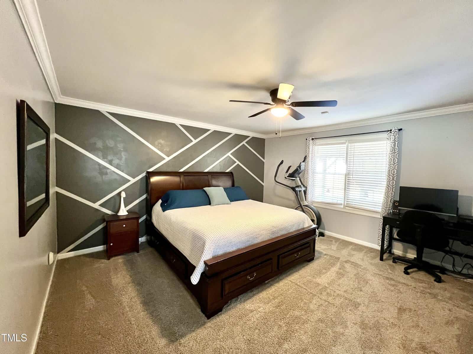 A bedroom with a large bed featuring a wooden frame and white bedding. A geometric accent wall is behind the bed. There's a ceiling fan, an exercise bike, a window with curtains, and a desk with a computer in the corner. Carpeted flooring completes the room.