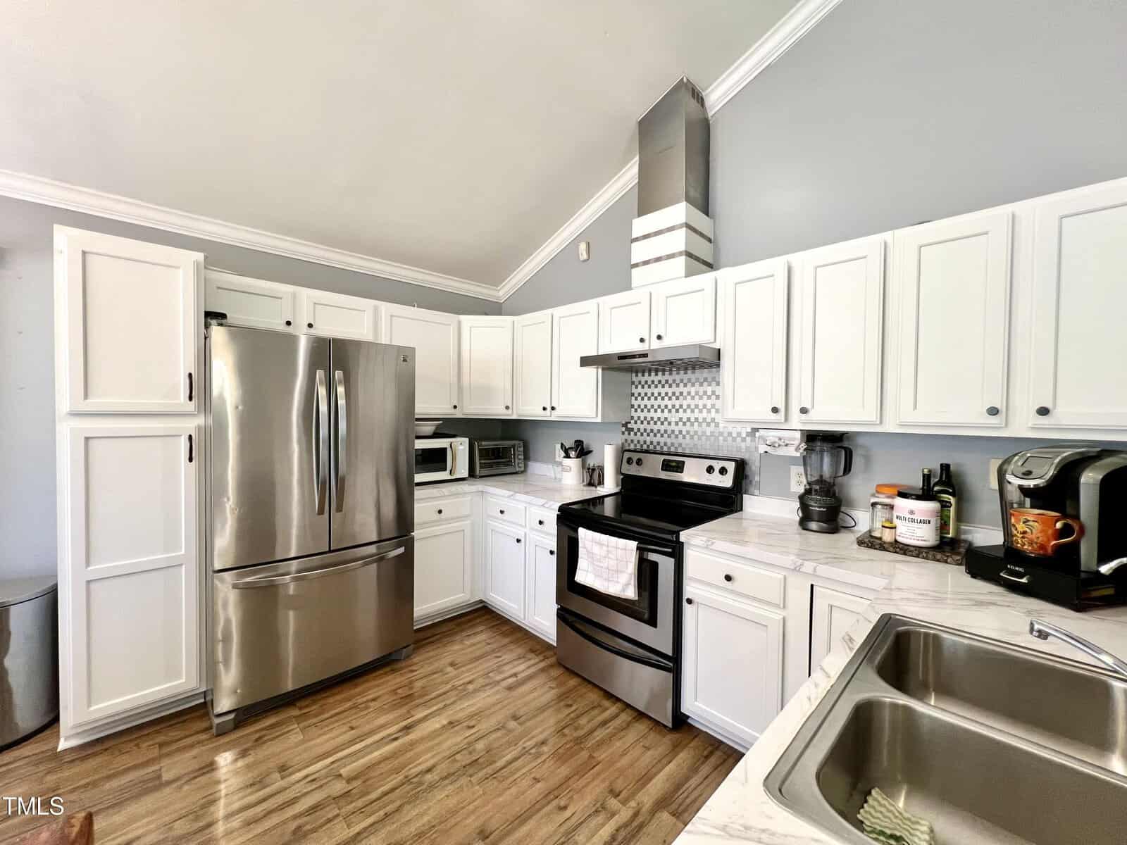 A modern kitchen with white cabinets and stainless steel appliances, including a fridge, oven, and microwave. The countertops are light-colored, with a coffee maker and a blender. A tile backsplash is visible behind the stove. The floor is wooden.