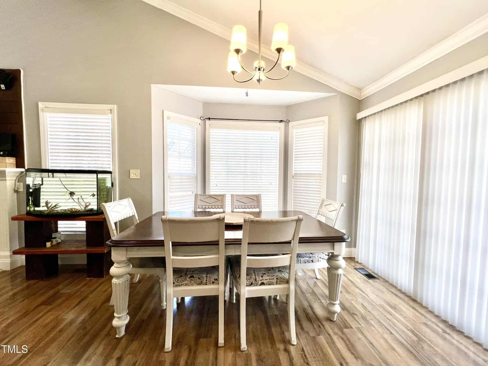 A bright, modern dining area with a wooden table and six white chairs, positioned near large windows with blinds. A chandelier hangs above the table. Light wood flooring and neutral-colored walls create a warm, inviting atmosphere.