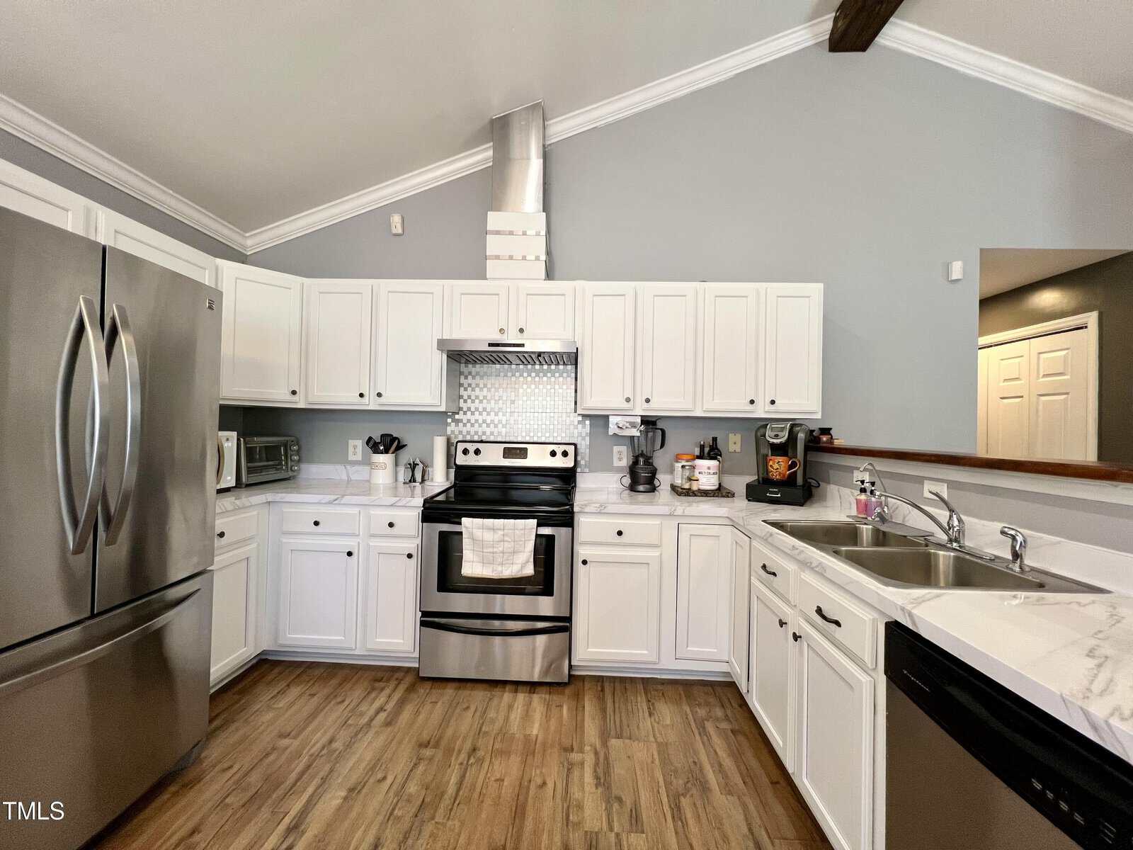 A modern kitchen featuring white cabinets, stainless steel appliances, and a wooden floor. There's a refrigerator on the left, an oven with a vent hood in the center, and a sink to the right with a view into another room.