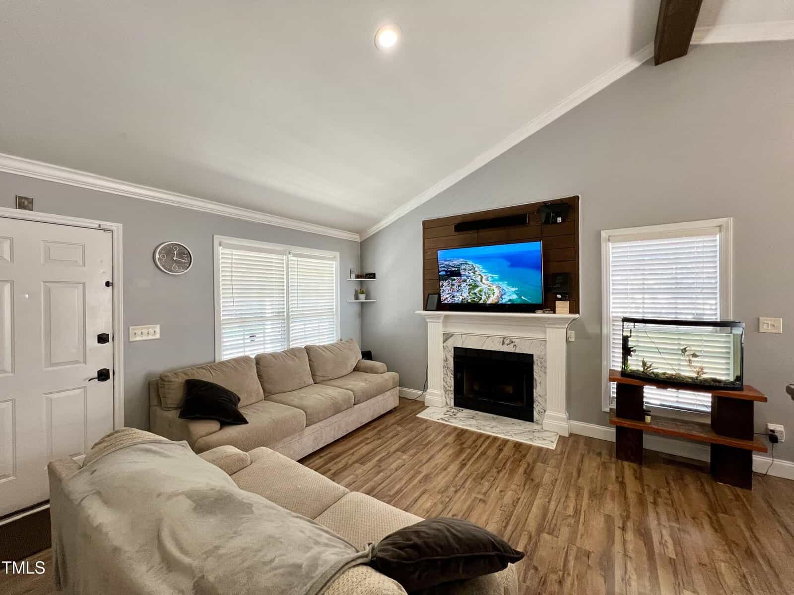 A cozy living room featuring two beige sofas, a wall-mounted TV above a fireplace, and a fish tank on a stand. The room has wooden floors, two windows with blinds, and a vaulted ceiling with a single recessed light.