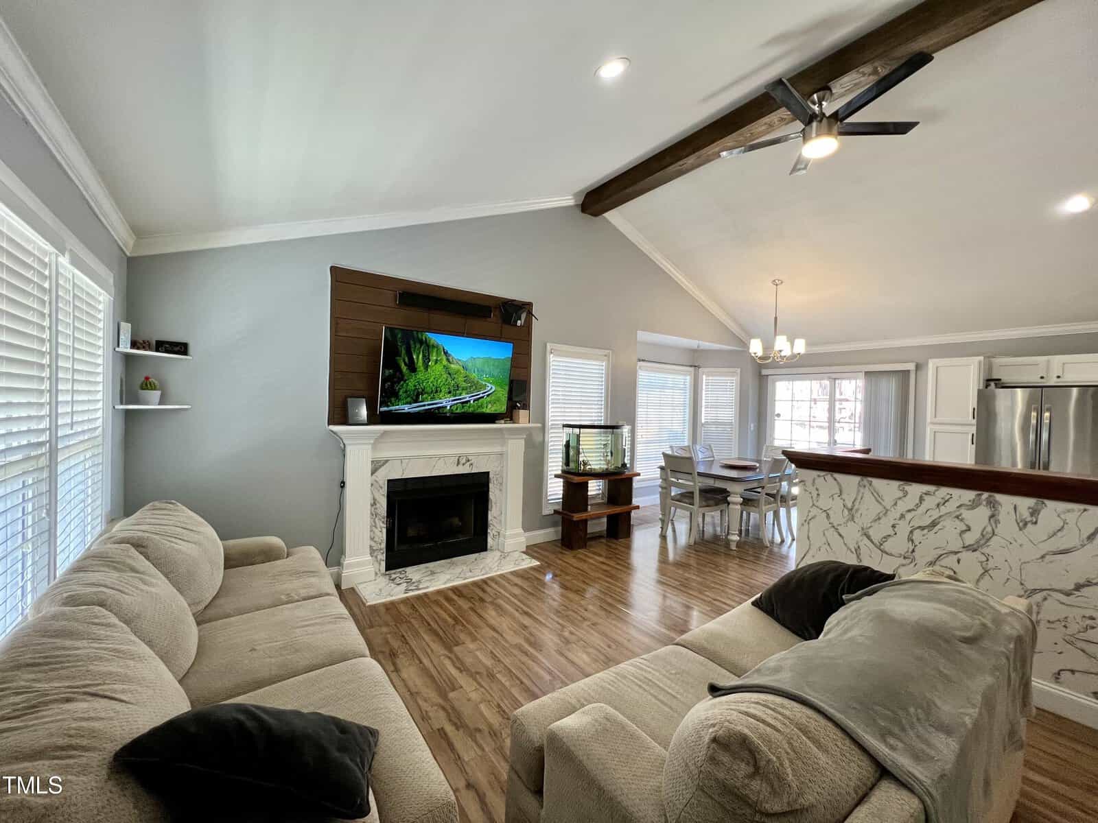 A cozy living room with two sofas facing a fireplace and a mounted TV. The room features a vaulted ceiling with exposed wooden beams, hardwood floors, and a kitchen island. A dining table and large windows provide ample natural light.