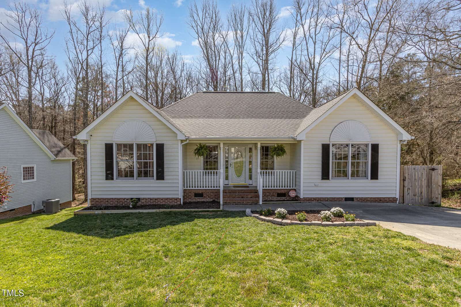 A charming single-story white house at 1608 Rogers Pointe Lane features a gable roof and brick foundation. With potted plants adorning the small porch and two symmetrical windows, this Creedmoor home is surrounded by a grassy lawn and leafless trees.