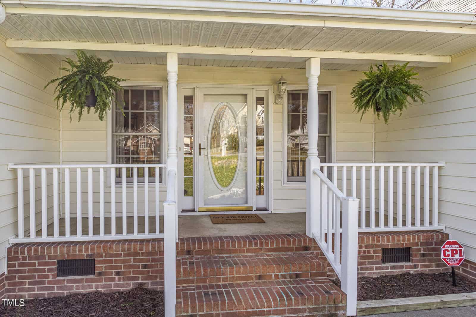 A front porch of a house with red brick steps leading to a white door with an oval glass panel. Two ferns hang from the ceiling on either side. A small red security sign is visible on the right side.