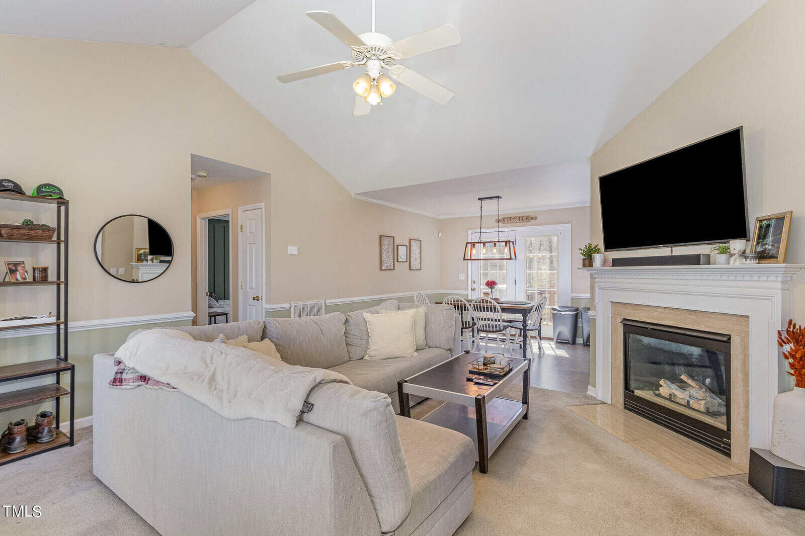 A cozy living room with a large beige sectional sofa, a coffee table, and a flat-screen TV above a fireplace. A ceiling fan hangs from a vaulted ceiling. The room features neutral decor, a round mirror, and a dining area in the background.