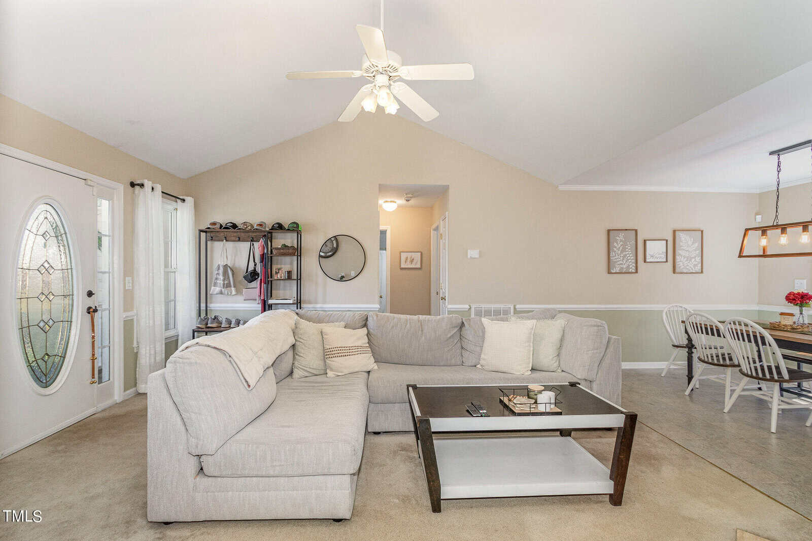 A cozy living room with a light gray sectional sofa, a wooden coffee table, and a ceiling fan. The open-plan area includes a dining space with a table and chairs. Neutral walls feature decorative elements, and a glass-paneled door is visible.