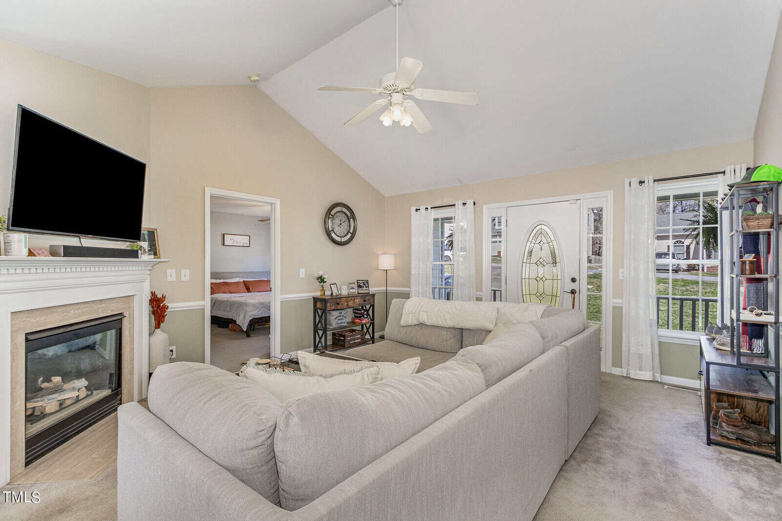 A cozy living room with a gray sectional sofa facing a television above a fireplace. An open door leads to a bedroom on the left. Large windows and a glass front door allow natural light. Neutral-colored walls and ceiling fan complete the space.