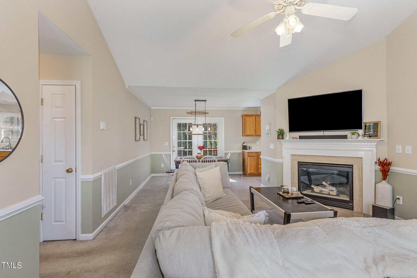 Living room with beige walls and carpet, featuring a large white sectional sofa. A fireplace with a TV above is on the right, and a dining area with a wooden table is in the background. Ceiling fan and decorative mirror visible.