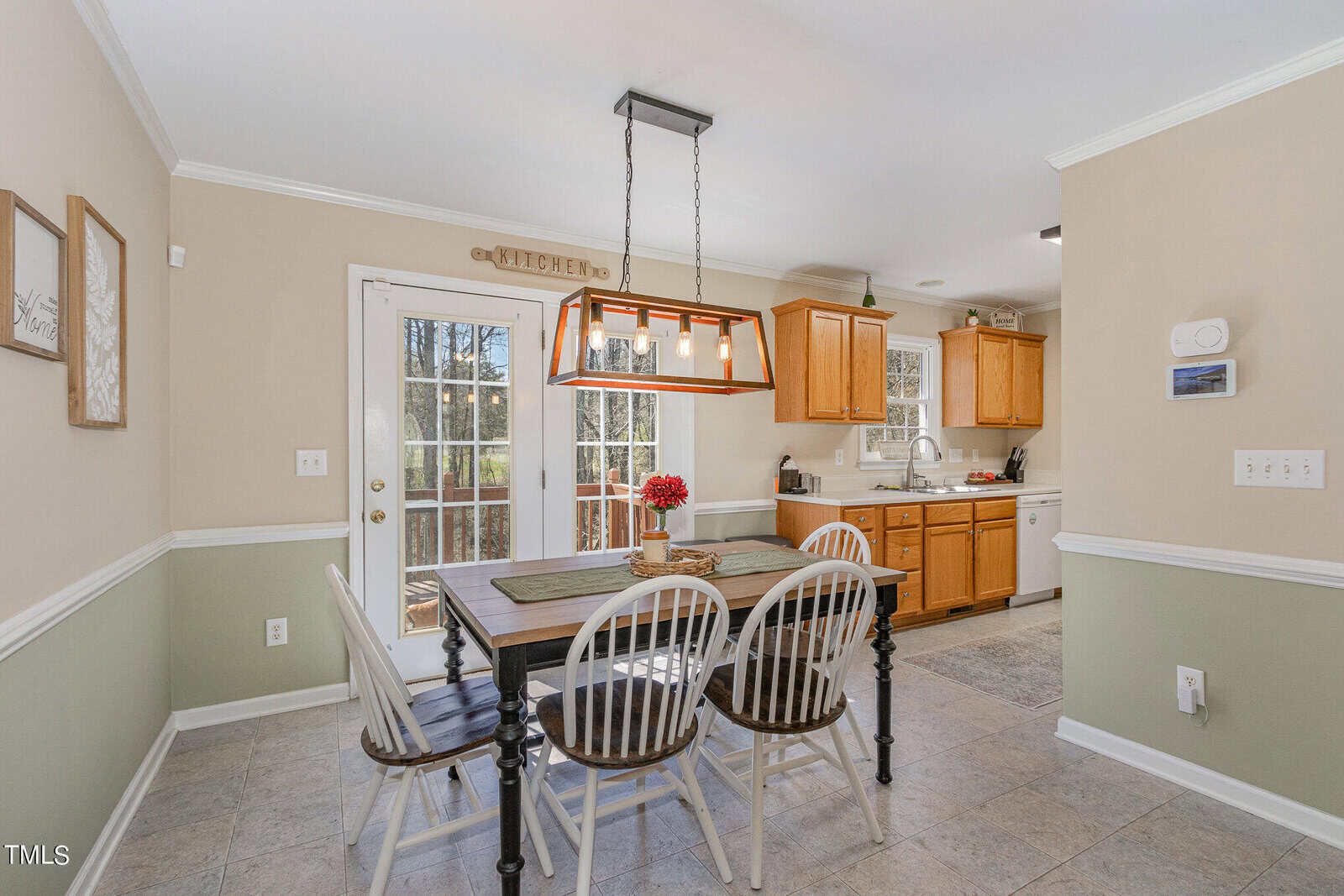 A cozy dining area with a wooden table and four chairs under a rectangular light fixture. French doors lead to the outside, and the kitchen with wooden cabinets is visible in the background. A "kitchen" sign and decor adorn the walls.