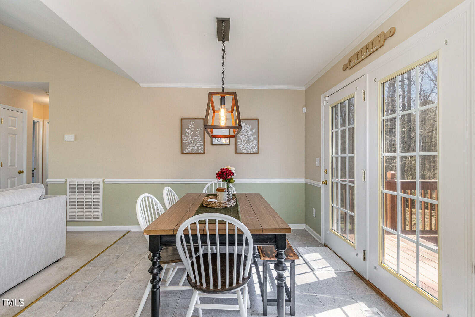 A dining room with a wooden table and four chairs, illuminated by a pendant light. Two wall hangings decorate the back wall. French doors on the right lead to a wooden deck. The room has beige walls and tile flooring.