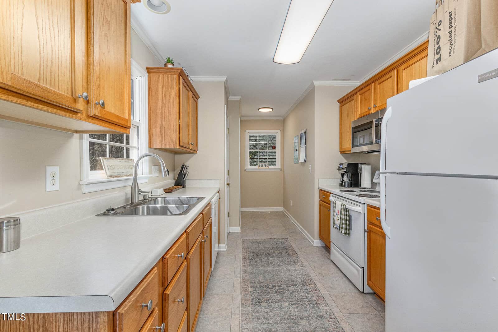 A narrow kitchen with wooden cabinets, white countertops, and a white fridge. A stove with a microwave above it is on the right, and a window on the left provides natural light. A small rug runs along the tiled floor.