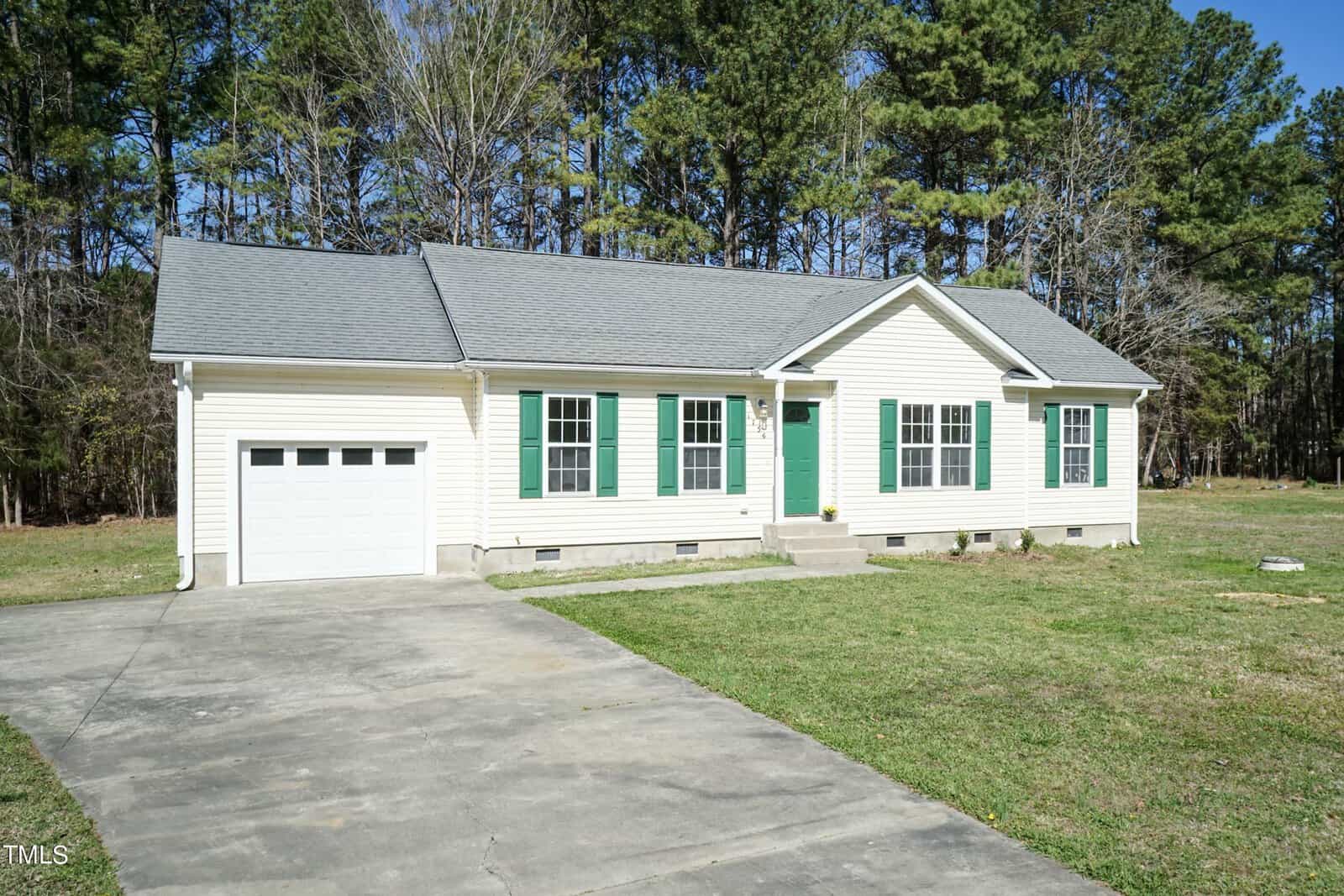 A charming one-story house at 1756 Cobblestone Drive, with beige siding and green shutters, nestles against tall trees. This Creedmoor home boasts a single-car garage with a concrete driveway leading up to it. The lawn is impeccably maintained under the clear blue sky.