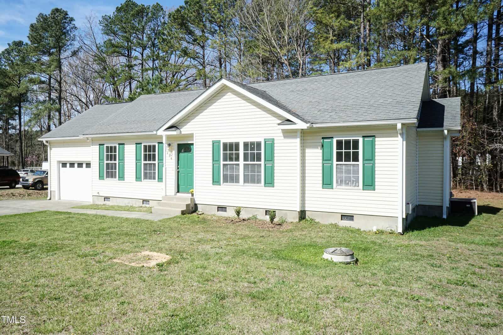 A single-story house with light beige siding and green shutters. It has a gray roof, a small front porch, and a one-car garage. The house is surrounded by a grassy lawn and bordered by tall trees.