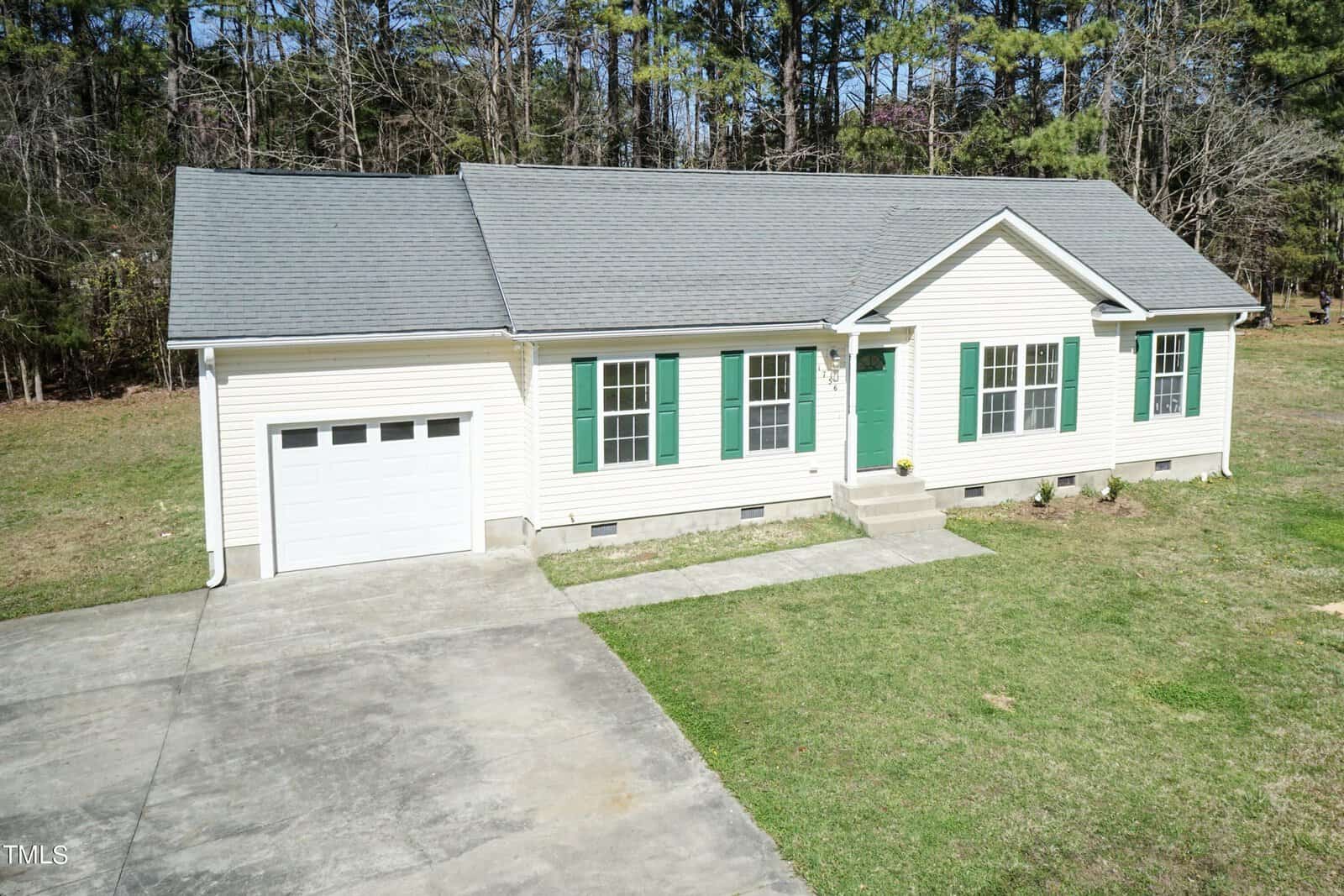 A single-story house with light yellow siding, green shutters, and a green front door. It has a one-car garage and a concrete driveway. The house is surrounded by a lawn and bordered by tall trees in the background.