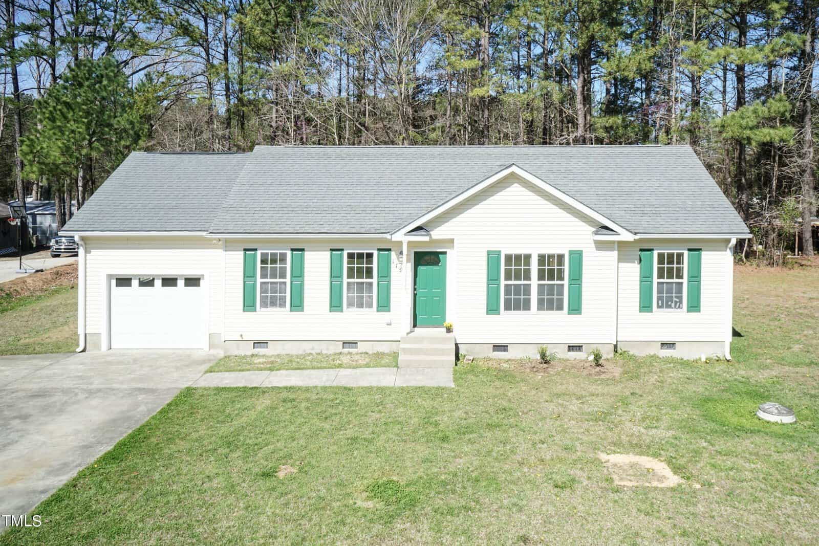 A single-story house with pale yellow siding and green shutters. It features a green front door, an attached garage, and a small front step. The house is surrounded by a grassy yard and trees in the background.