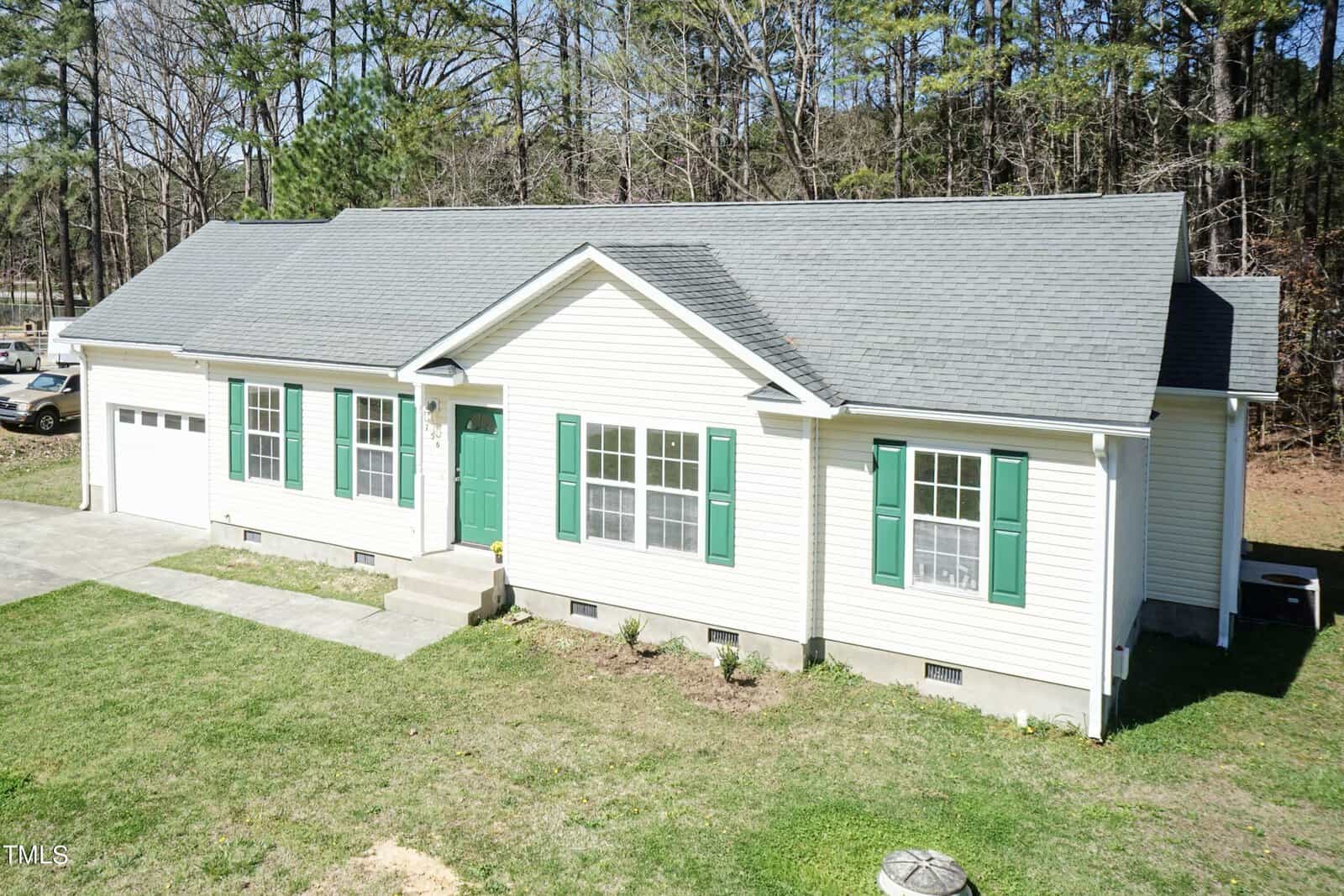 A single-story white house with green shutters and a gray roof. It has a small front porch, a two-car garage on the left, and is surrounded by grass and trees in the background.