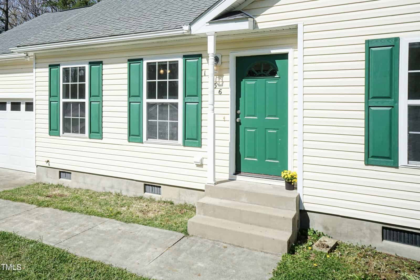 A beige house with green shutters and a matching green front door. A small porch leads to the entrance, with a potted plant beside the door. A concrete pathway is in front of the house, and the garage is partially visible.