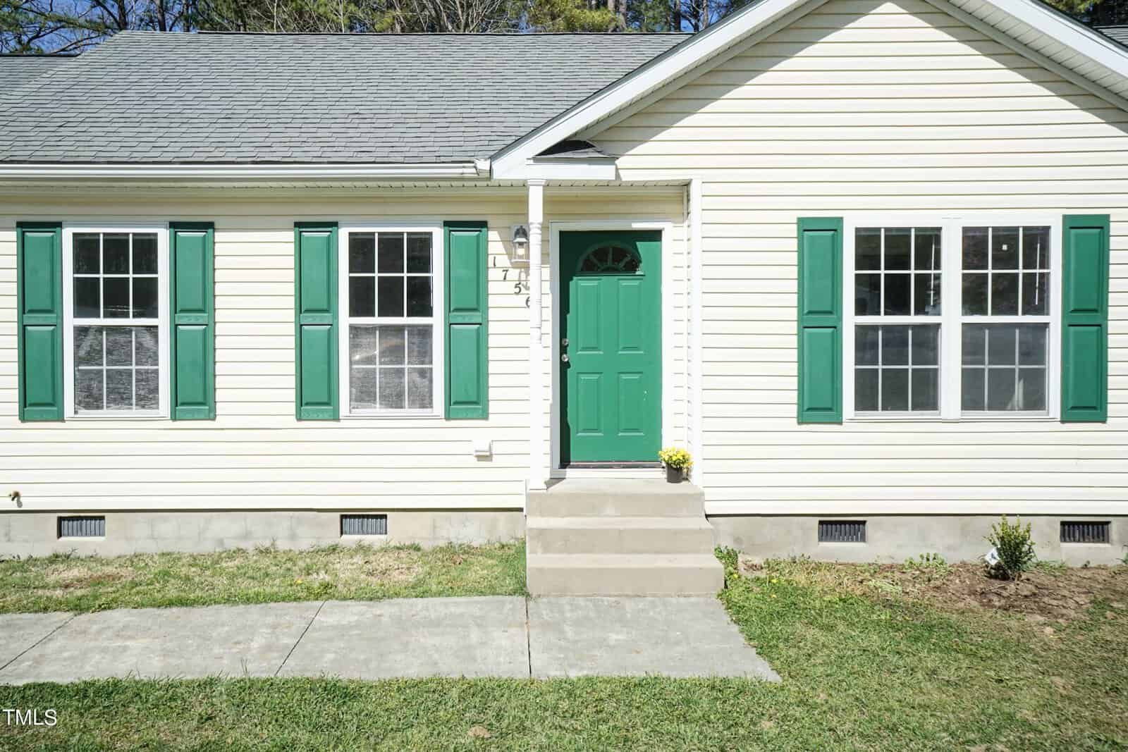 A beige house with green shutters and a green front door. There are three windows and steps leading up to the entrance, with a small plant next to the door. A walkway is visible in the front yard.