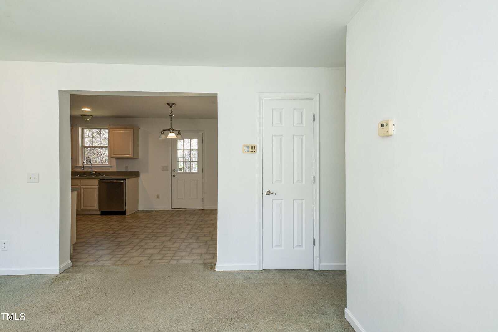 A minimalist interior showing an open space with a white door leading to another room. The area has white walls, gray carpet, and tiled flooring near the kitchen, which features white cabinets and a stainless steel dishwasher. A ceiling light hangs above.