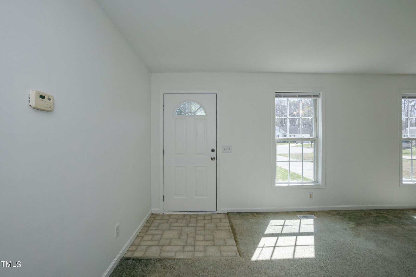 A bright, empty room with white walls and carpeted floor. There is a white door with a small window and two large windows to the right. Sunlight streams through, casting light on the floor.