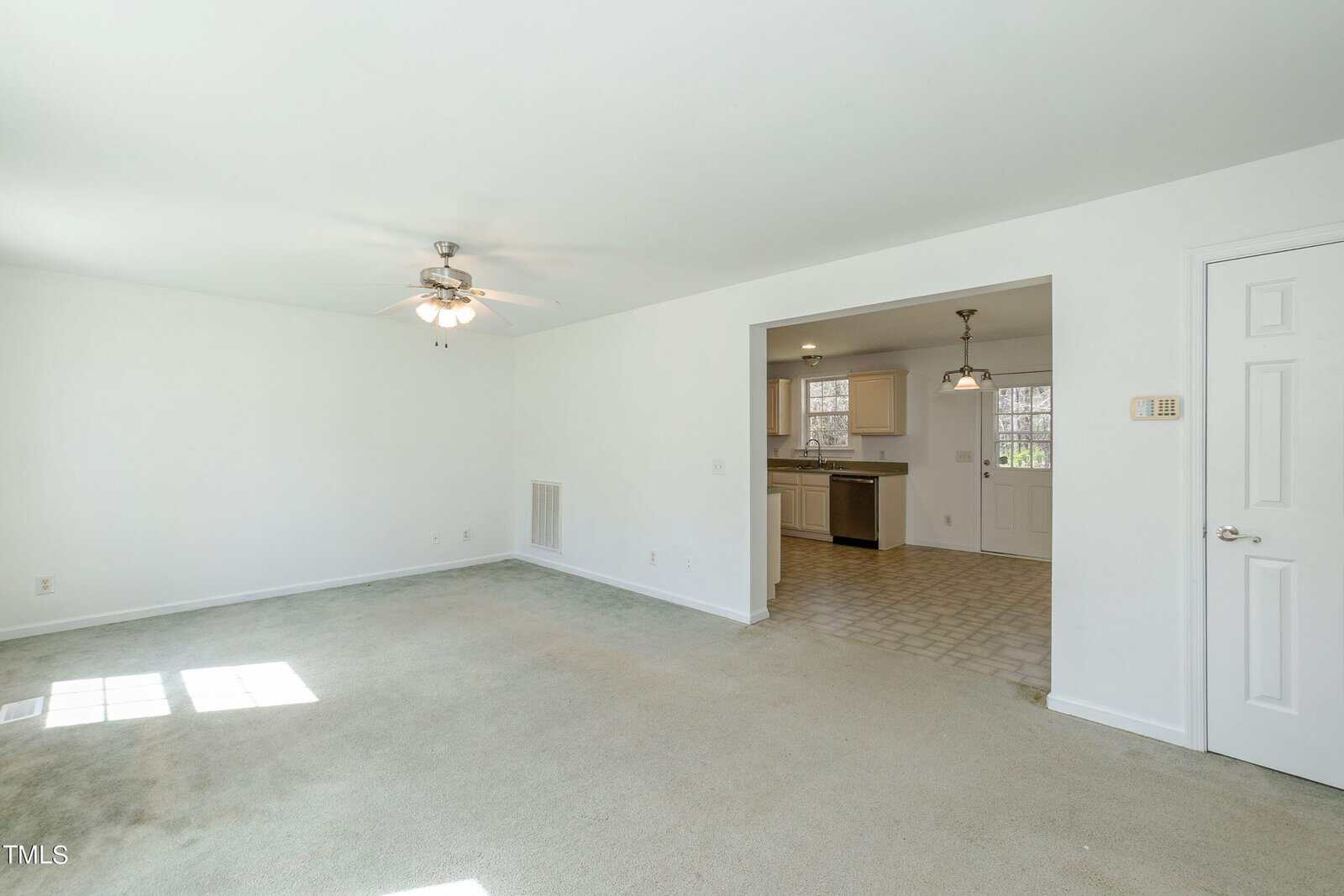 A spacious, empty living room with light carpet and white walls. There's a ceiling fan and a large open doorway leading to a kitchen with white cabinets and tile flooring. Sunlight pours in through a window, creating a bright, airy atmosphere.