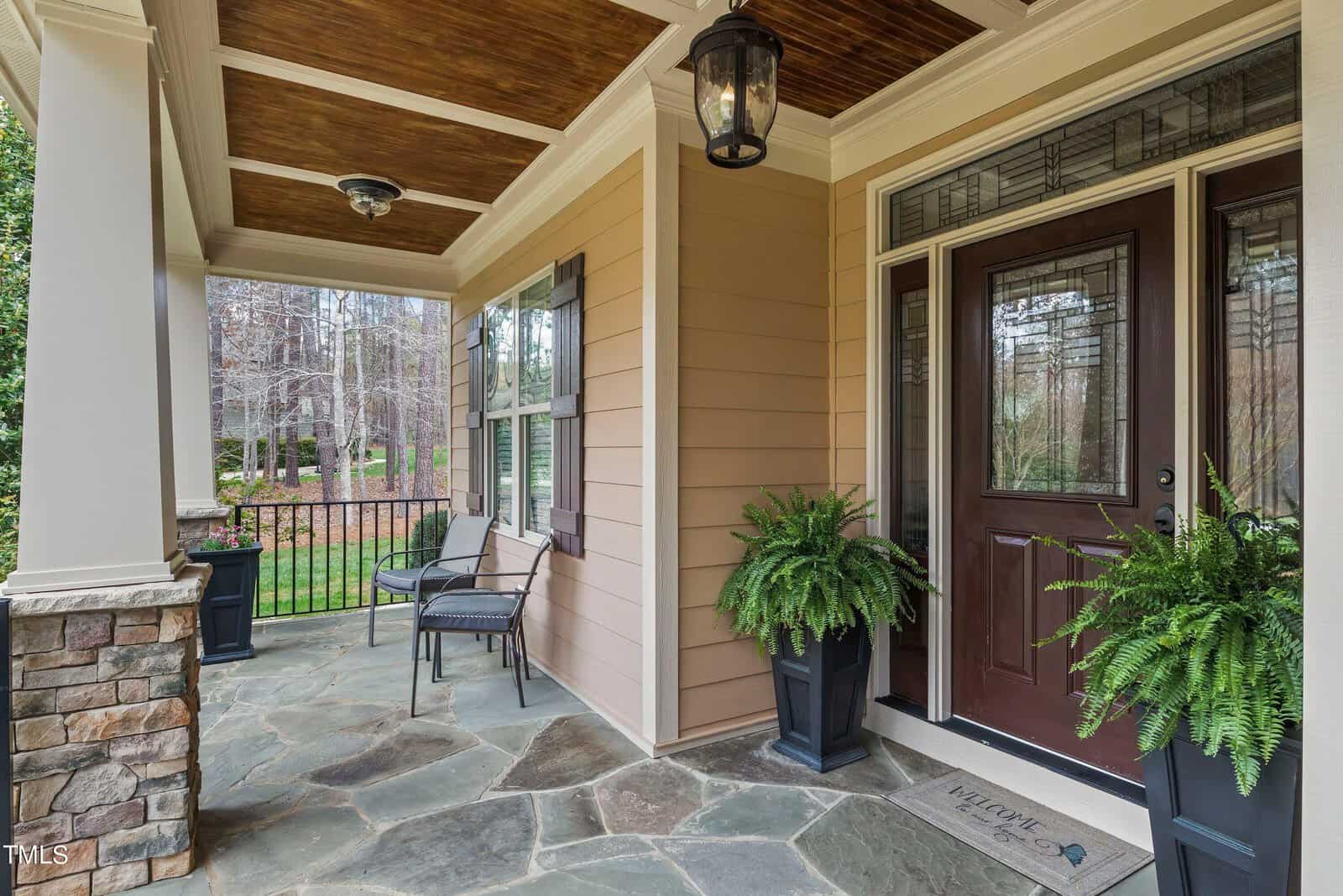 A cozy porch at 3584 Cotton Field Court features a wooden ceiling and stone flooring. The dark wooden door with decorative glass panels is flanked by two potted ferns, accompanied by a seating area with black chairs. A welcome mat invites visitors to this Wake Forest home, with trees visible in the background.