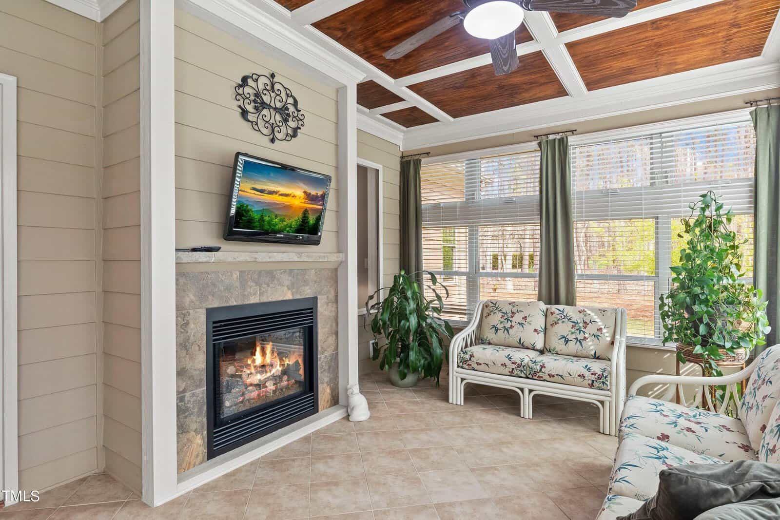 A cozy sunroom features a stone fireplace with a TV mounted above it, surrounded by wicker furniture with floral cushions. Large windows allow natural light, and green plants add a touch of nature. The ceiling has wooden panels and a fan.