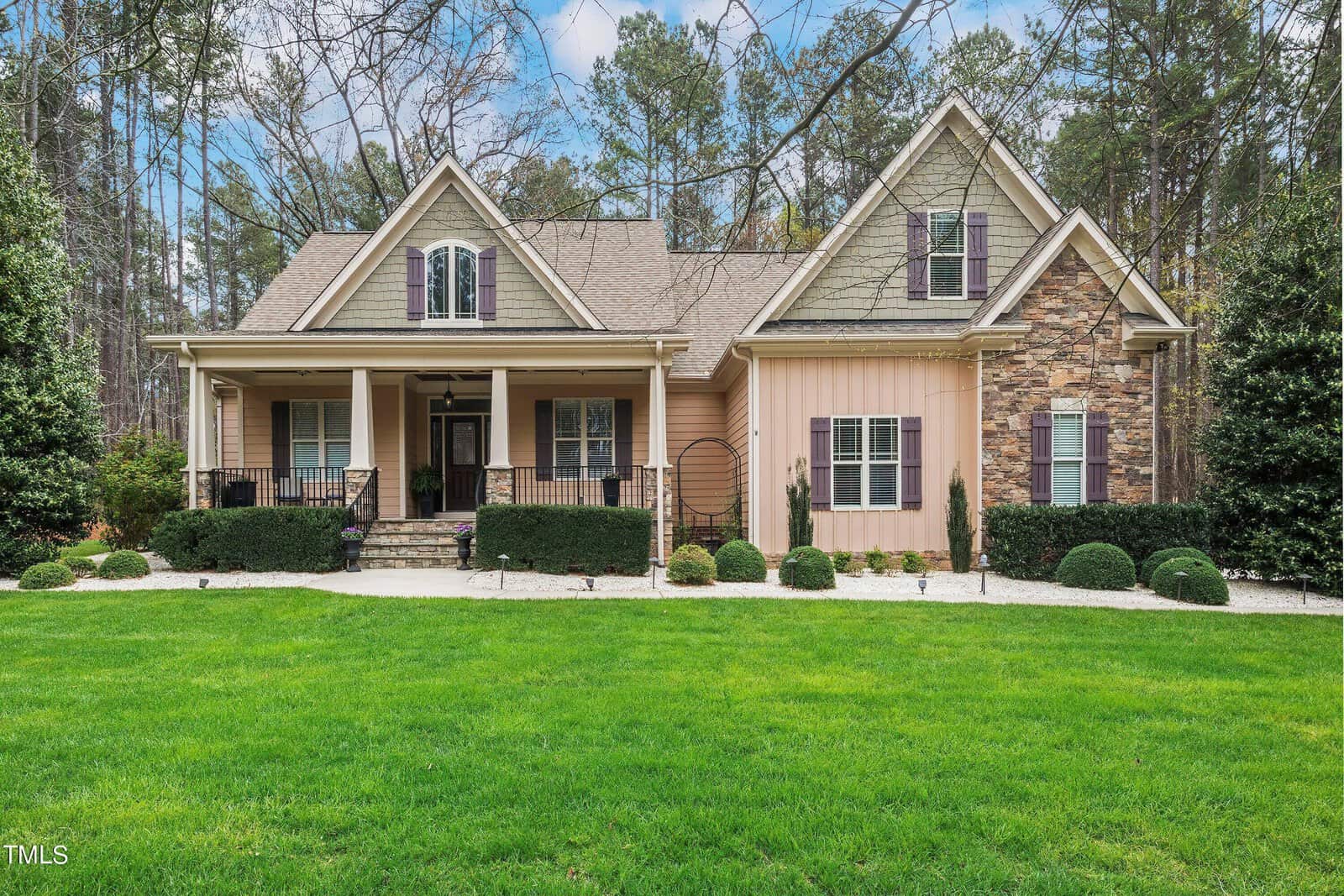 A charming suburban house with a symmetrical facade, featuring a stone and siding exterior, a front porch with columns, and a well-maintained grassy yard. Tall trees are visible in the background under a partly cloudy sky.