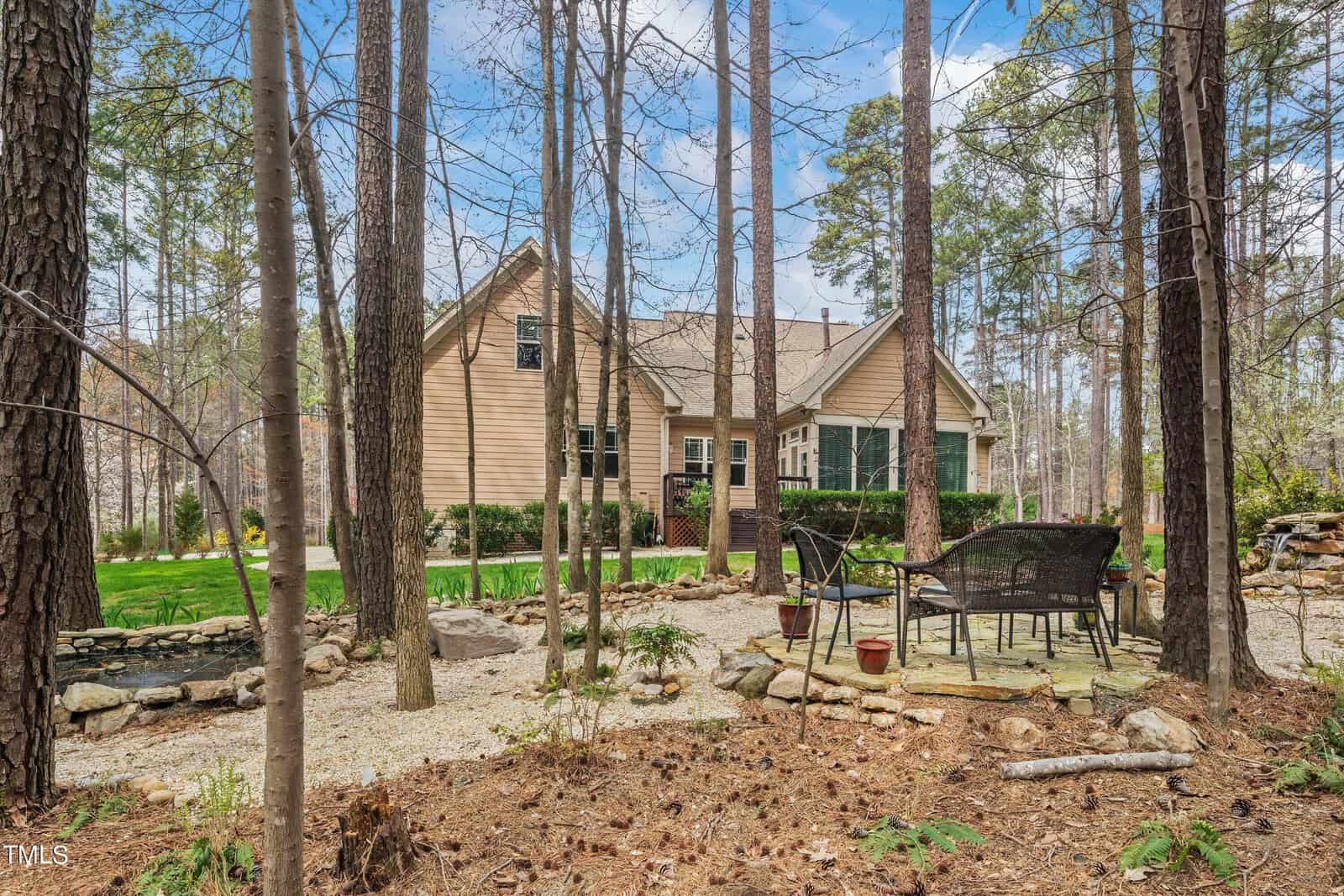 A cozy wooden house surrounded by tall trees. In the foreground, there is a patio area with chairs and a small table on a gravel surface. Lush greenery and stone accents enhance the natural setting.