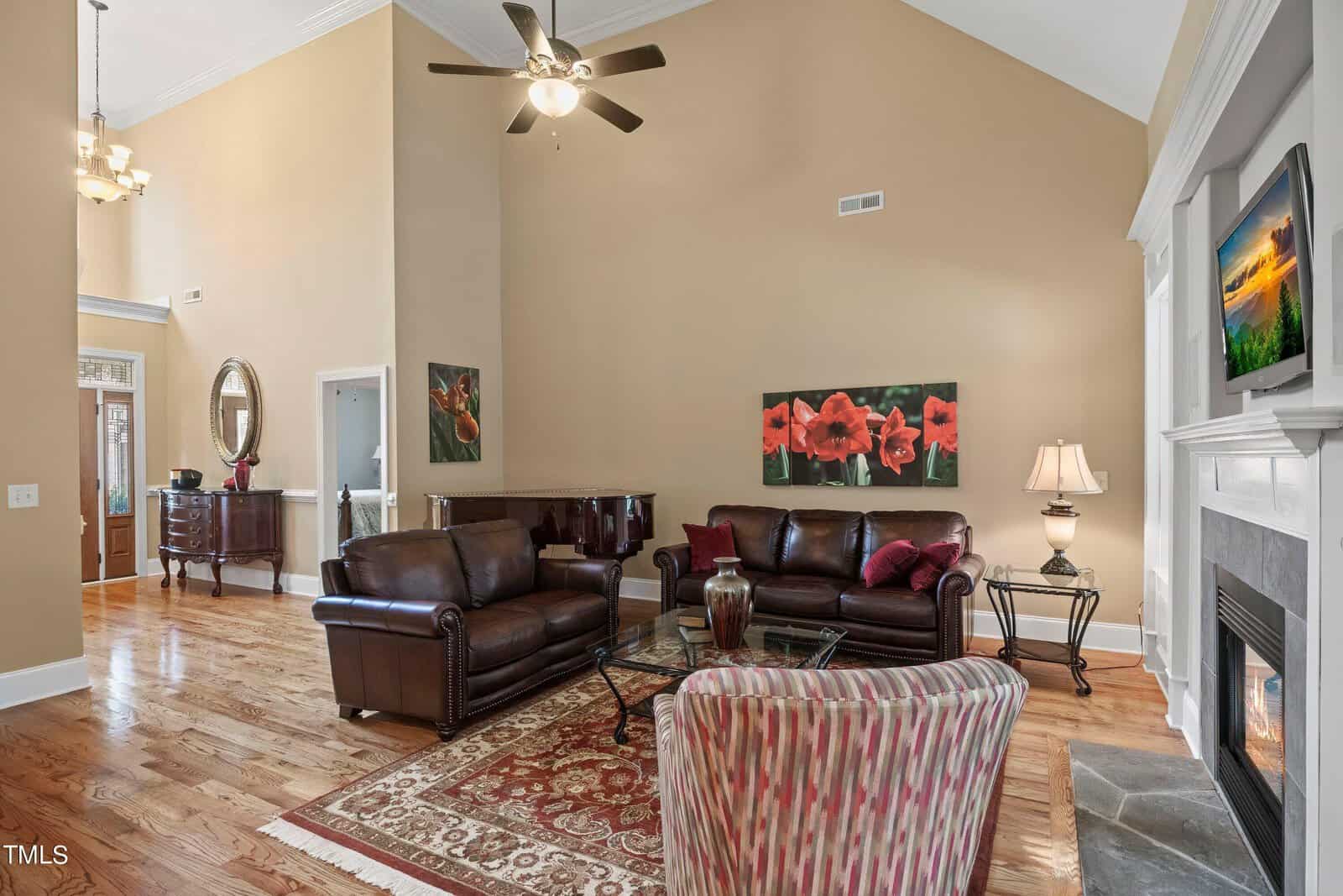 A spacious living room with high ceilings and hardwood floors. It features a leather sofa set, a glass coffee table, and a striped armchair. There are red floral paintings on the wall, a fireplace, and a ceiling fan with lights.