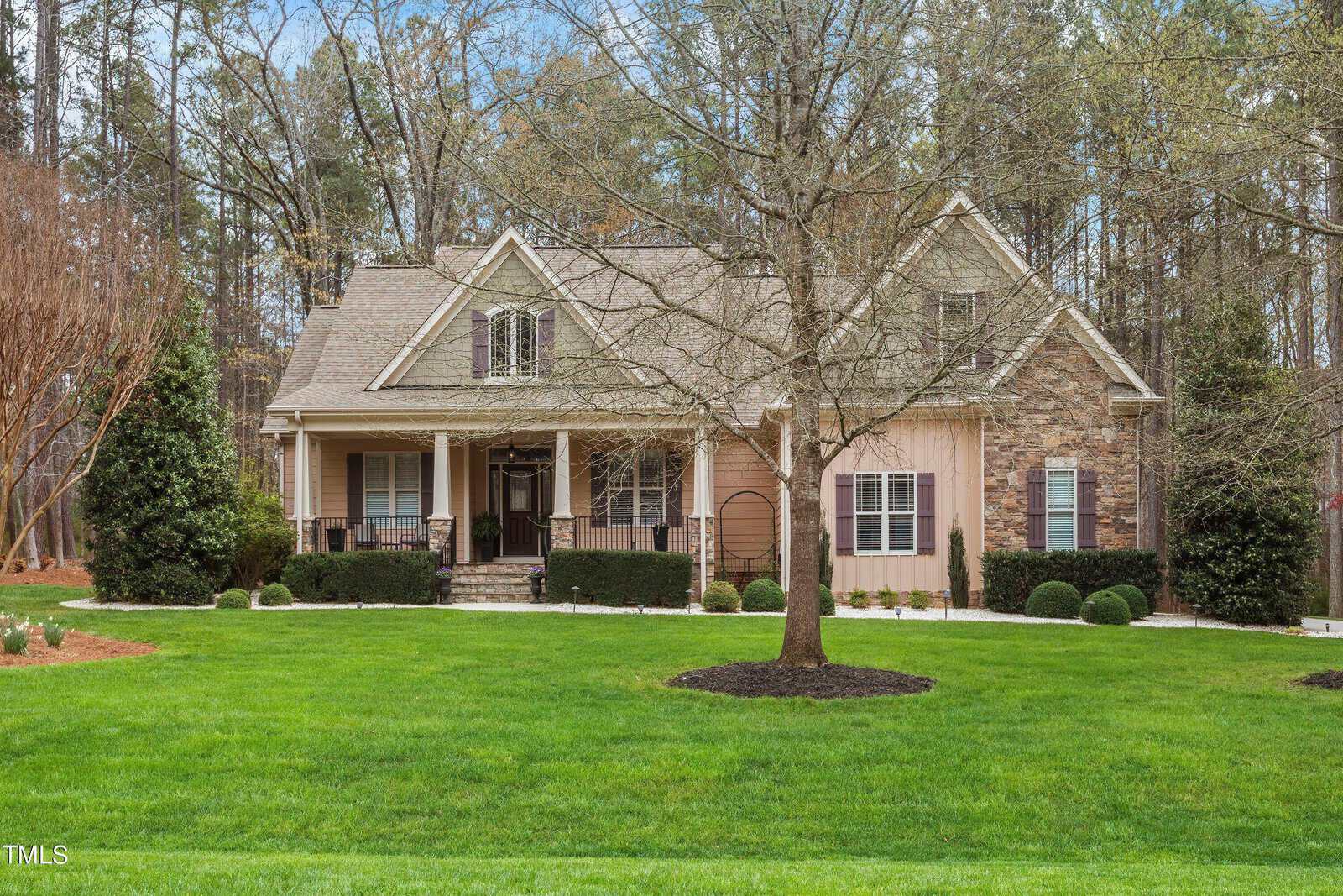 A two-story house with a stone and wood exterior, featuring a gabled roof and a covered porch. The front yard is landscaped with a large tree, shrubs, and a well-maintained lawn, surrounded by tall trees in the background.