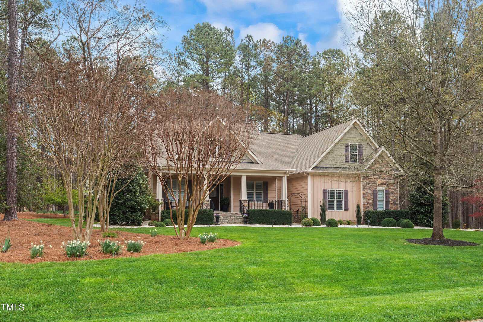 A charming suburban house with stone and wood exterior, surrounded by a well-kept lawn and trees. A pathway leads to the front entrance, and there are flower beds with blooming plants. The sky is partly cloudy with some blue patches.