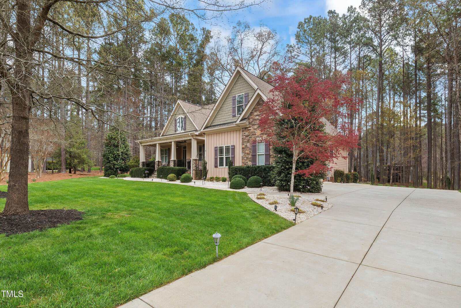 A charming house with a large front porch and manicured lawn set against a backdrop of tall trees. The driveway curves gracefully, bordered by neat shrubbery and a tree with red foliage, all under a partly cloudy sky.