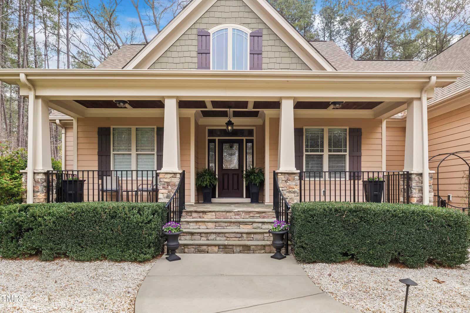 A charming suburban house with a covered porch featuring stone accents and two potted plants. It has beige siding, a dark front door, and two windows with shutters. A pathway leads to the entrance, surrounded by manicured bushes and gravel.