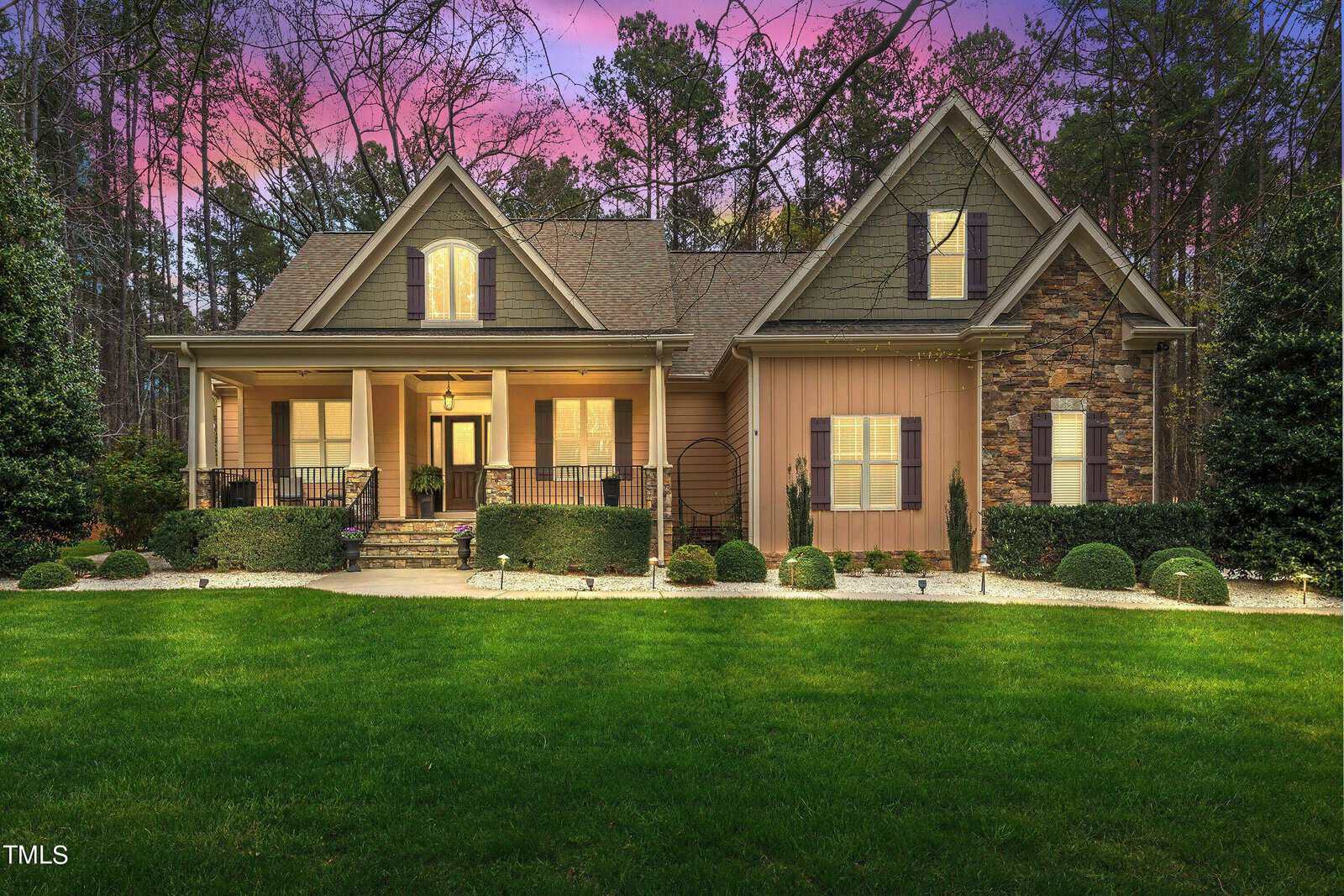 A charming house with tan siding and stone accents, featuring a well-maintained lawn and front porch. Surrounded by tall trees, the sky is a vibrant mix of pink, purple, and blue during sunset.
