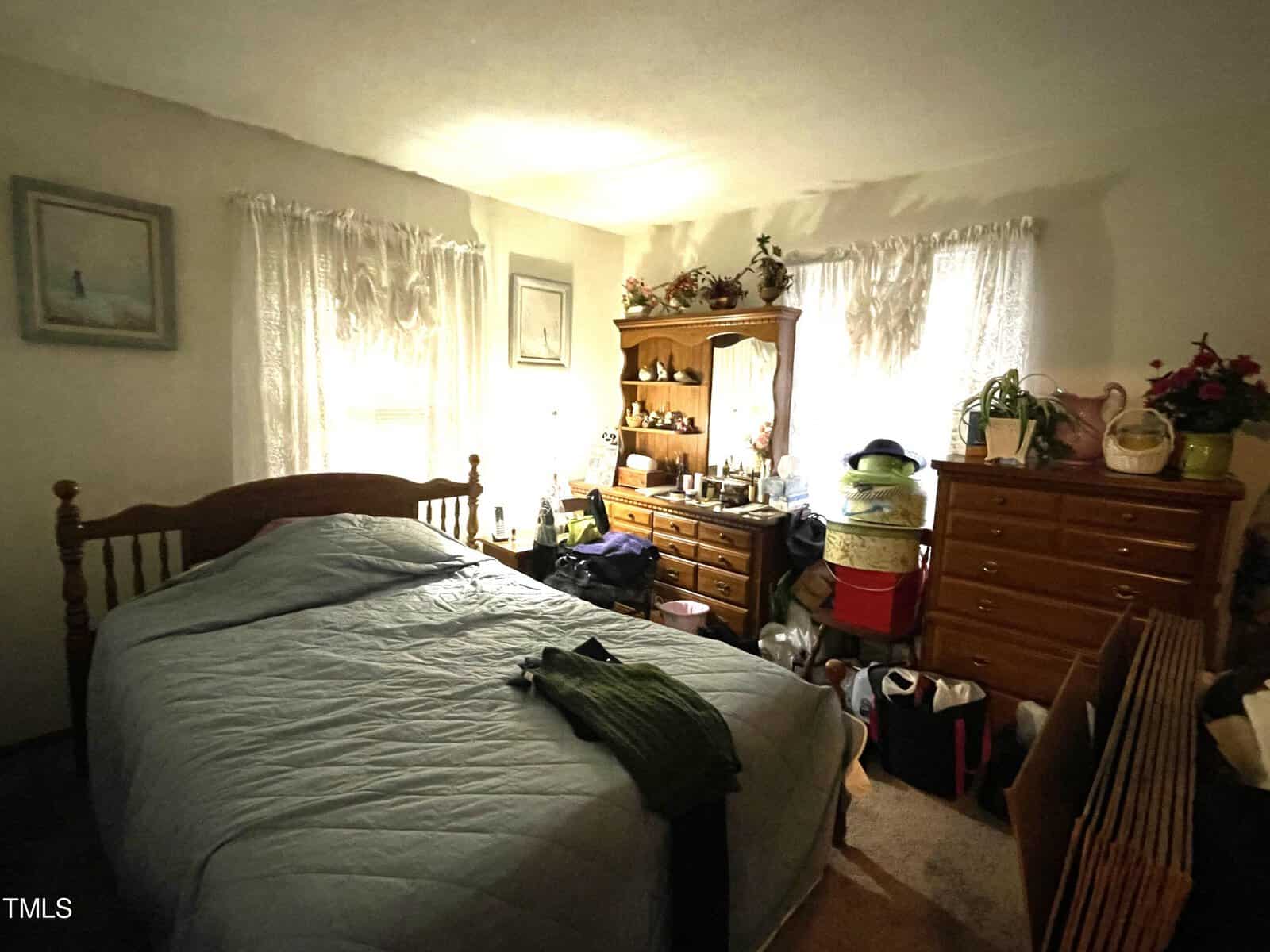A cozy and slightly cluttered bedroom with natural light filtering in through lace curtains, featuring a wooden bed, dresser, various personal items, and decorative plants.