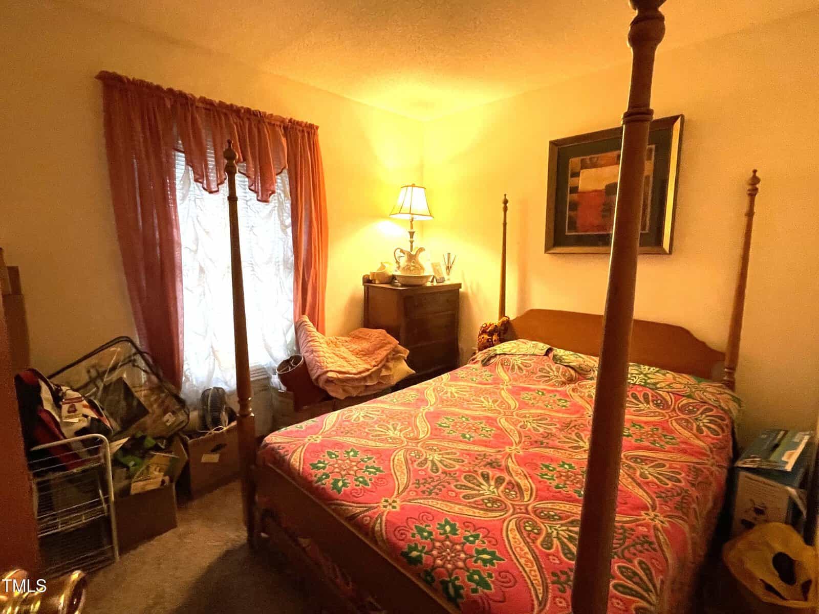 A cozy bedroom with a pink and green patterned bedspread on a four-poster bed, a cluttered side area, a wooden dresser with a lamp, and a window dressed in long red curtains.