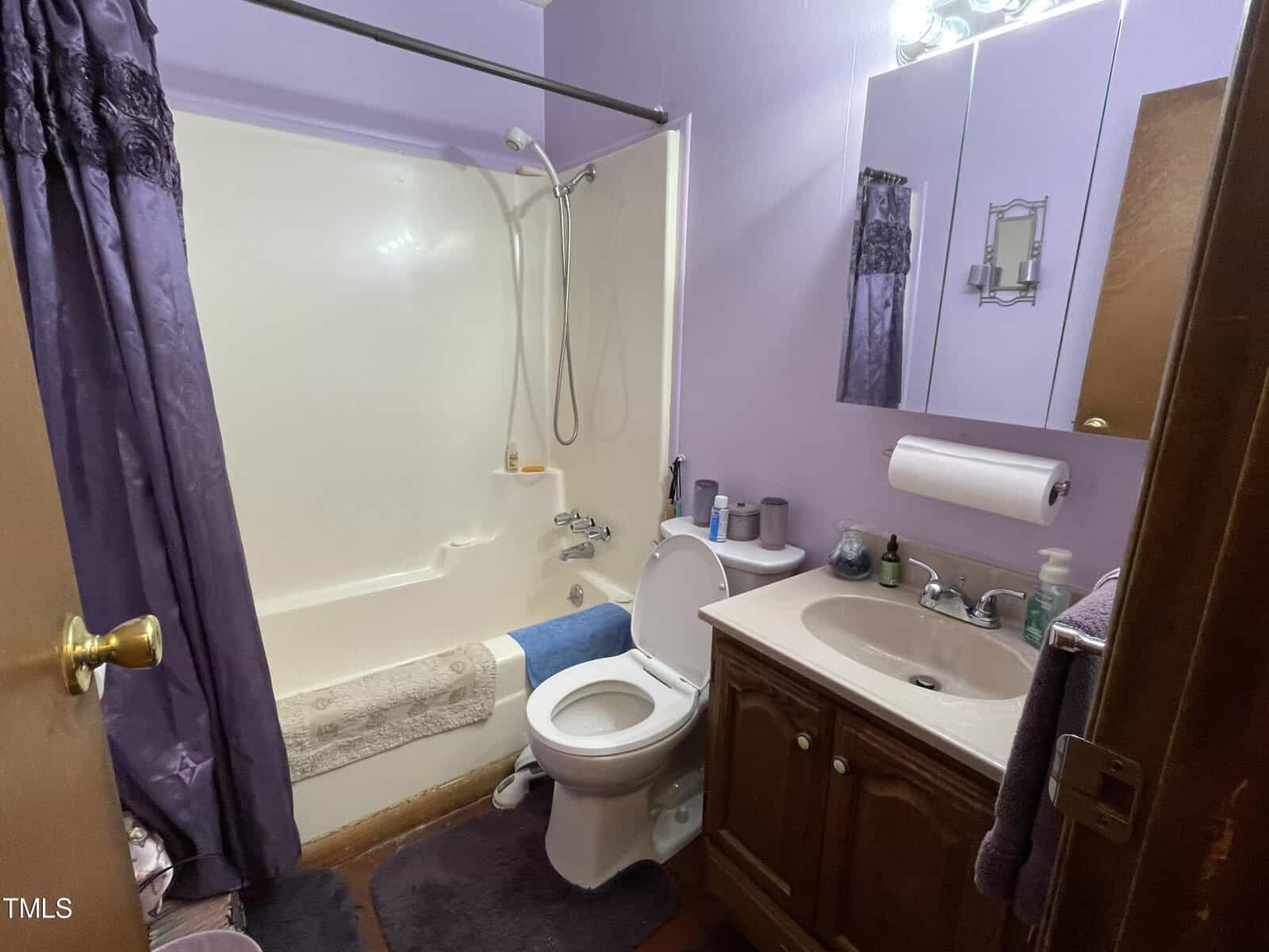 A small bathroom with a shower-tub combo, toilet, and sink vanity. the room has purple walls, a white curtain, and various toiletries.